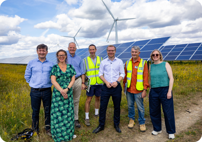 Michael Shanks MP, with a team from Westmill in front of solar panels and turbines with a blue cloudy sky and green grass behind them. Credit: DESNZ