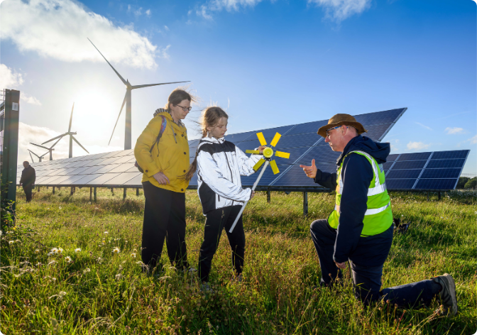 Child with adult next to her, holding a yellow model wind turbine, with a man in a hi vis jacket knelt down next to her. Solar panels and wind turbines can be seen in background, with a blue sky and bright sun shining.